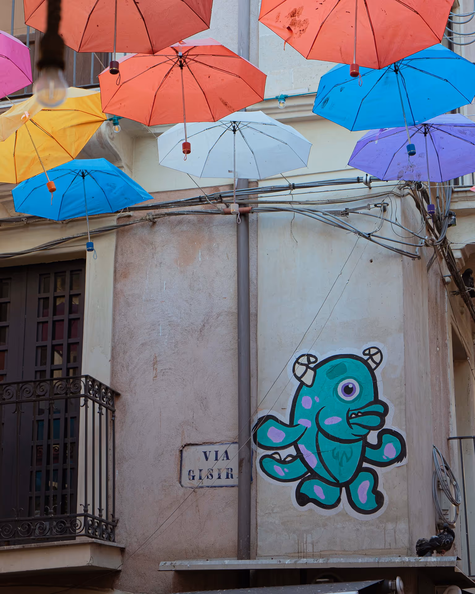 Street art of a teal monster, with bright coloured umbrellas hanging above