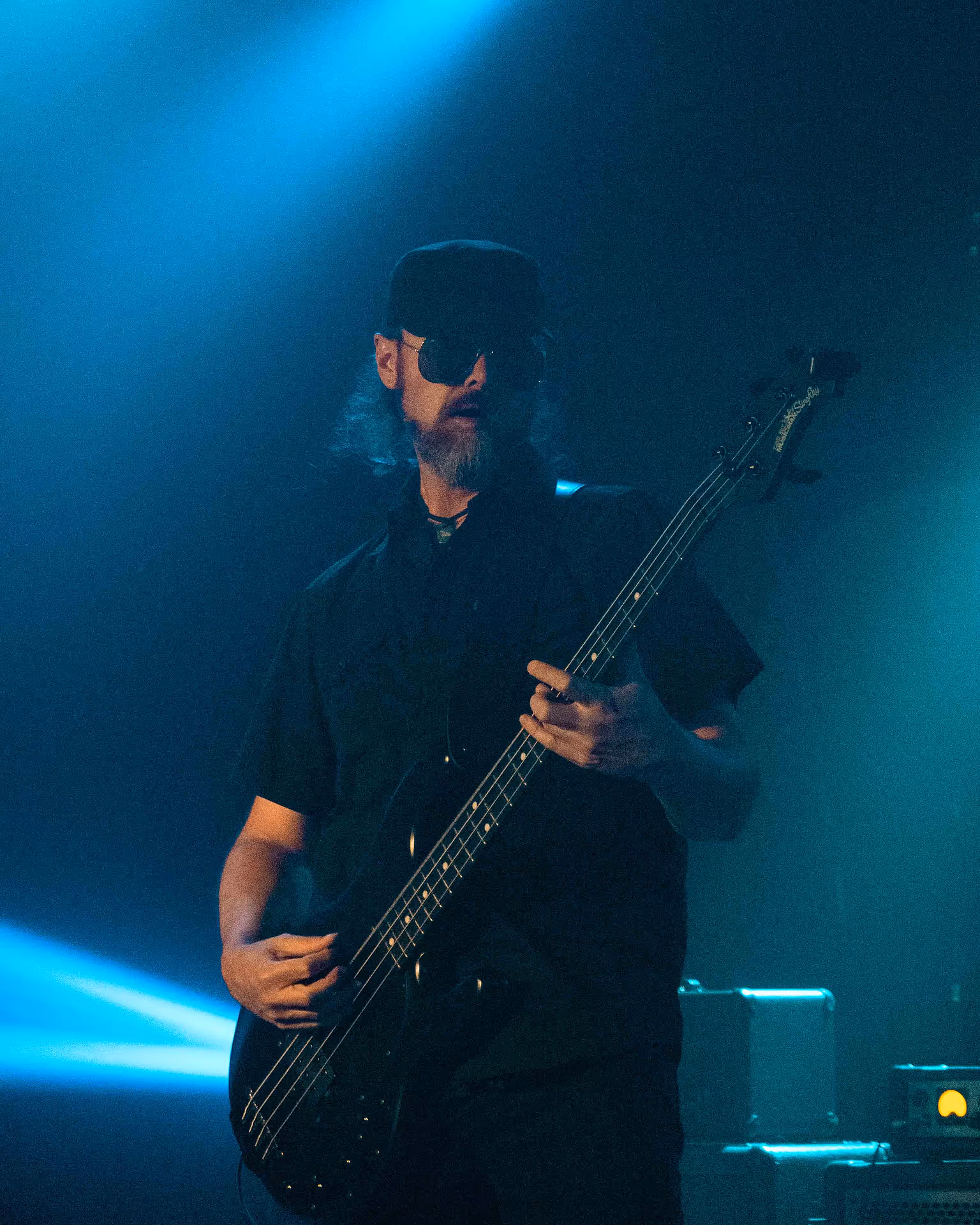 Man playing a bass guitar, wearing sunglasses with blue concert lights.